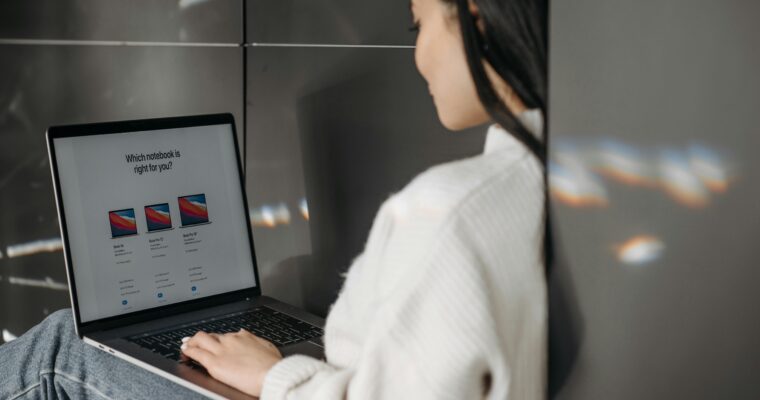A woman in a white sweater sitting on the floor against a wall on a laptop.