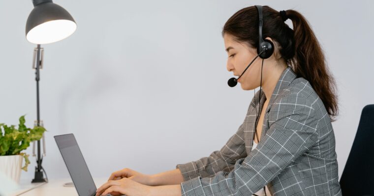 A woman sitting at a desk working on a laptop under a desk lamp and wearing a headset.